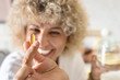 Close-up of a cheerful, curly-haired woman displaying a golden Omega-3 capsule to the camera with a blurry background, symbolizing health, wellness, and dietary supplements at home.