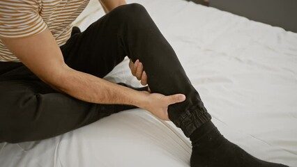 Wall Mural - Hispanic man in casual attire sitting on a white bed at home touching his leg, indicating pain or discomfort.