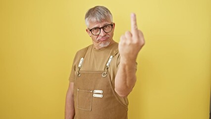 Canvas Print - Impudent middle-age grey-haired man in glasses and apron standing, showcasing a rude 'fuck you' hand gesture. angry expression over isolated yellow wall background