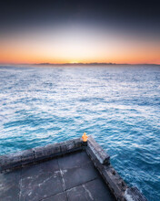 Aerial View Of A Man In An Orange Jacket, Standing On The Edge Of A Jetty Looking Out Towards The Sunset With Mountains On The Horizon, Kalk Bay, Western Cape, South Africa.