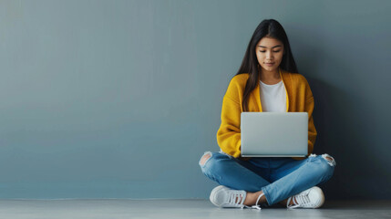 Canvas Print - A young woman in a yellow sweater and jeans sits on the floor with her legs crossed, engrossed in her laptop.