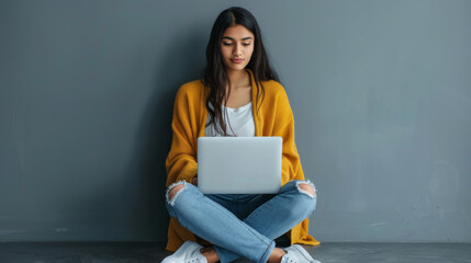 Canvas Print - A young woman in a yellow sweater and jeans sits on the floor with her legs crossed, engrossed in her laptop.