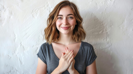 Sticker - A contented young woman in a casual grey shirt holds her hands together in a gesture of gratitude, smiling gently against a textured white wall.