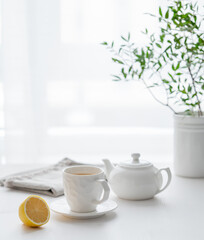 Wall Mural - A cup of tea with lemon, a teapot and green branch on a white table against the background of a kitchen window.