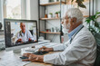 An experienced elderly male doctor with white hair and glasses, dressed in a professional white lab coat with a stethoscope around his neck, is engaged in a telemedicine consultation.