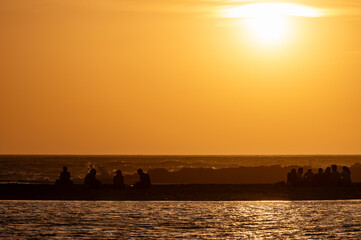 Group of men and women sitting and looking at sunset on ocean beach, orange sky, silhouettes of people on vacation