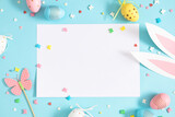Fototapeta Tulipany - Easter party concept.  Flat lay, top view of blank sheet of paper, easter bunny ears white pink blue and yellow eggs, colorful sugar candies on isolated pastel blue background