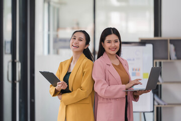 Wall Mural - Two beautiful Asian businesswoman golding digital tablet and laptop, standing in the office room.