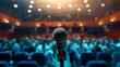 A single microphone on a stand is highlighted by a spotlight against a blurred background of an auditorium filled with an expectant audience, suggesting a live performance or speech.