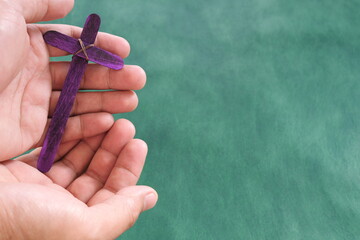 Wall Mural - Male hand holding purple wooden cross flat lay in green background. Holy week and lent season celebration concept.