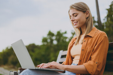 Wall Mural - Side view young happy IT woman wears orange shirt casual clothes hold use work on laptop pc computer sit on bench walk rest relax in spring green city park outdoors on nature. Urban lifestyle concept.