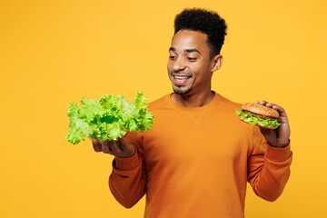 Wall Mural - Young fun man wear orange sweatshirt casual clothes hold lettuce salad burger choose what to eat isolated on plain yellow background studio Proper nutrition healthy fast food unhealthy choice concept