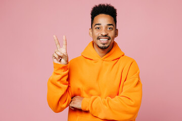 Poster - Young smiling happy man of African American ethnicity wear yellow hoody casual clothes show victory sign look camera isolated on plain pastel light pink background studio portrait. Lifestyle concept.
