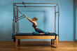 © Georgii - A young girl does Pilates exercises with a bed reformer, barrel machine tool. Beautiful slim fitness trainer on the background of a reformer doing various exercises gray, low key, light art. Fitness