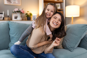 Poster - Happy Beautiful Mother carrying or piggyback her little daughter laughing playing and having fun together on sofa at home.