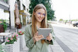© muse studio - Urban professional woman using a tablet, smiling on a city street with flowers.