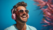 Joyful young man with stylish sunglasses and pink headphones enjoying music, against a vibrant blue background