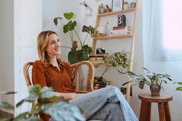 Portrait of a young cheerful woman sitting on the chair in her living room early in the morning and looking at the window. Copy space, interior background.