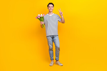 Canvas Print - Full length photo of optimistic cheerful guy wear stylish t-shirt jeans pants hold tulips show v-sign isolated on yellow color background