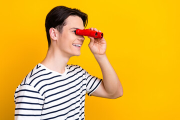 Sticker - Portrait of positive guy with brunet hair wear stylish t-shirt look at empty space through binoculars isolated on yellow color background