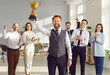 © Studio Romantic - Happy winner businessman receives a business award. Joyful man holding his trophy, with a team of cheerful workers in the background celebrating and congratulating their leader on great work success