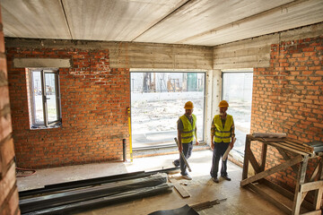 two hardworking men in safety vests working on site with blueprints and level, cottage builders