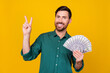 Photo of cheerful pleasant guy with beard dressed dotted shirt holding money showing v-sign symbol isolated on yellow color background