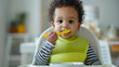 © Studio Nova - Toddler self-feeding with spoon in high chair.