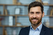 © Liubomir - Close-up photo of a portrait of a young successful businessman, lawyer, broker standing in a suit in the office, smiling and confidently looking at the camera