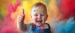 A smiling baby with blue eyes, wearing overalls, is covered in colorful paint smudges on the face and hands, giving a thumbs-up gesture, with a vibrant explosion of powder paint colors in the backgrou