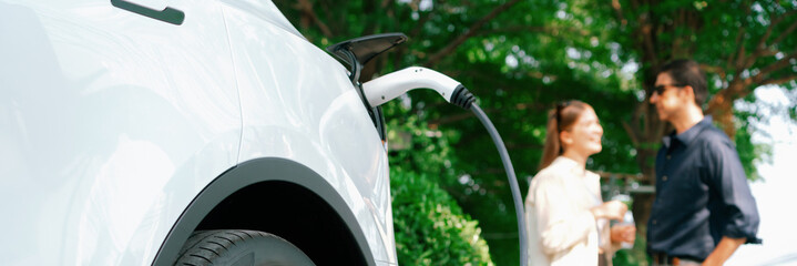 young couple recharge electric car battery from charging station in green city park in springtime. r