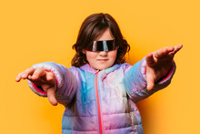Confident Girl Posing With Futuristic Glasses And Extending Her Hands Forward, Wearing A Colorful Pastel Jacket Against An Orange Background