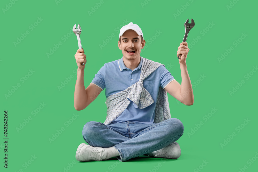 Young man with wrenches sitting on green background
