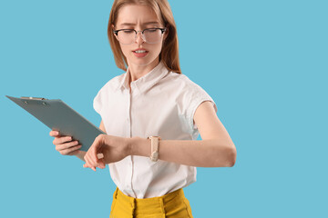 Wall Mural - Young businesswoman with clipboard looking at wristwatch on blue background. Time management concept