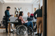 © qunica.com - A modern office setting with multicultural team members and a colleague in a wheelchair, emphasizing diversity and inclusion at work.