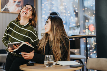 Two young women engaged in a cheerful discussion at work, with one holding a notebook in a warmly lit room decorated with twinkling lights.