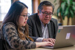 Mature Latin manager mentor talking to young Asian female coworker showing online project results at meeting. Two happy diverse professional executives team working in office using pc laptop