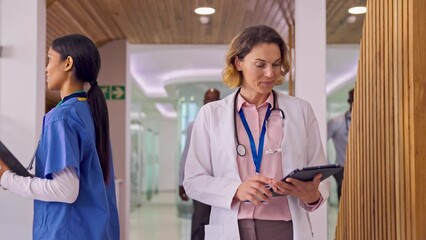 Wall Mural - Female Doctor In White Lab Coat With Digital Tablet Walking Past Colleagues In Hospital 