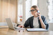 © muse studio - Multitasking businesswoman in glasses on a phone call while working on her laptop, in a bright office, a model of modern efficiency.