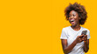 Mujer afro sorniente utilizando su smartphone. Chica negra sobre fondo amarillo. Persona utilizando tecnología con espacio de copia.