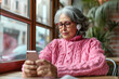 © wolfhound911 - Elderly gray haired Latin American female in glasses and pink sweater sitting at table adjusting earphones while listening to audio on mobile phone