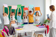 © Studio Romantic - Children with colourful sheets of paper in rising up hands on class with teacher in elementary school standing near the desk. Game form of work in lesson. Learning of colors using play activities.