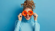 © MP Studio - A person holds bright orange gerbera flowers over their eyes like glasses, smiling broadly against a blue background, creating a playful and joyful portrait.