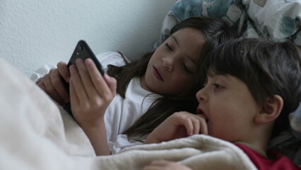 Wall Mural - Small siblings staring at cellphone device in bed, sister sharing phone screen with brother looking at smartphone device together