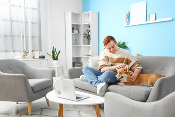 Sticker - Redhead young happy man with cute Corgi dog sitting on sofa at home