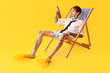 Tattooed young man with bottle of beer sitting on beach chair against yellow background