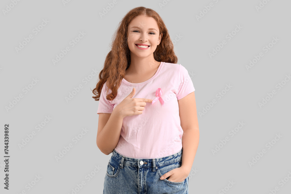 Young redhead woman pointing at pink ribbon on light background. Breast cancer awareness concept