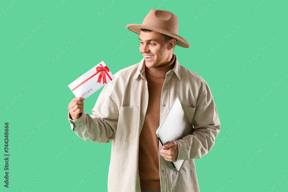 Young man with gift card and laptop on green background