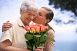 Senior woman, daughter and kiss with flowers, hug and care for love, bonding and reunion at family home. People, elderly mom and mothers day celebration with connection, gift and bouquet in garden