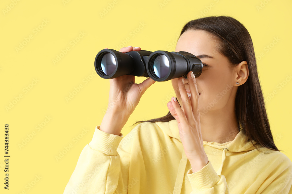 Young woman looking through binoculars on yellow background, closeup